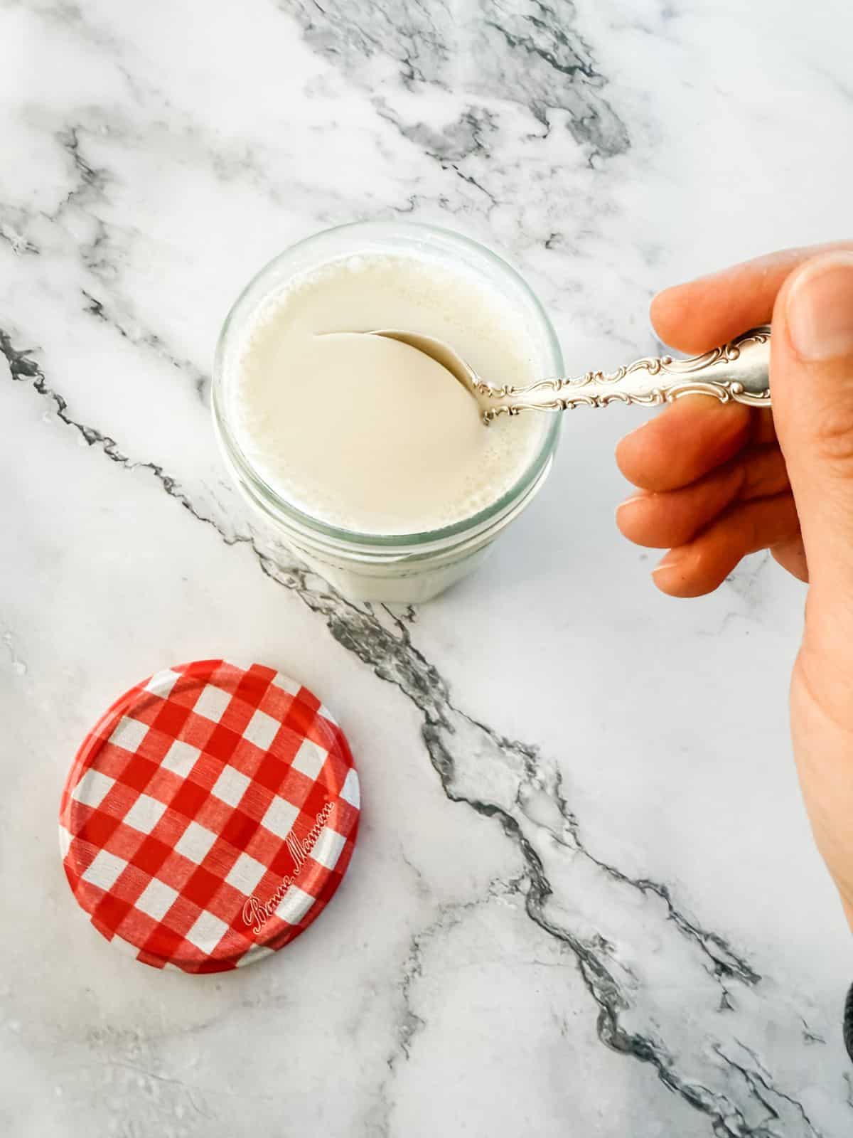 Homemade pot of yoghurt with spoon held by hand on white marble bench top. 