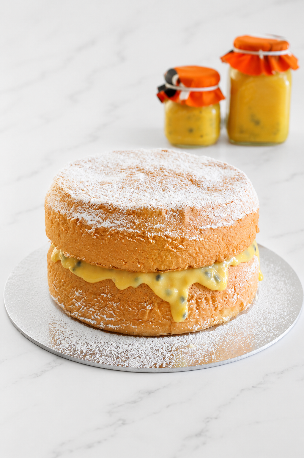 Double layer sponge cake sandwhiched with passionfruit curd on a silver plate on white marble bench top. 