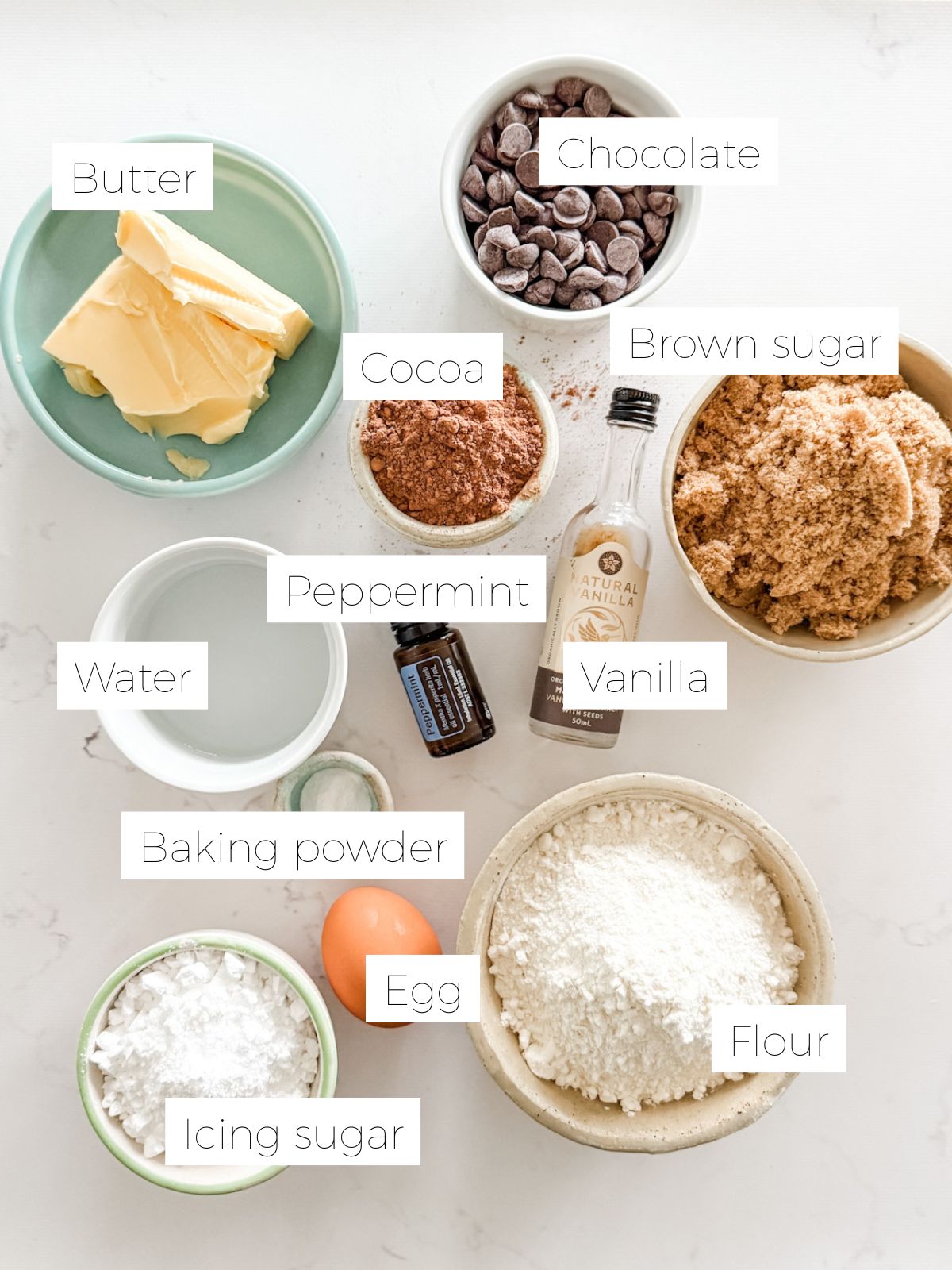 Butter, bowls of brown sugar, chocolate, flour, icing sugar, water, an egg, vanilla and peppermint oil to make choc mint biscuits. 