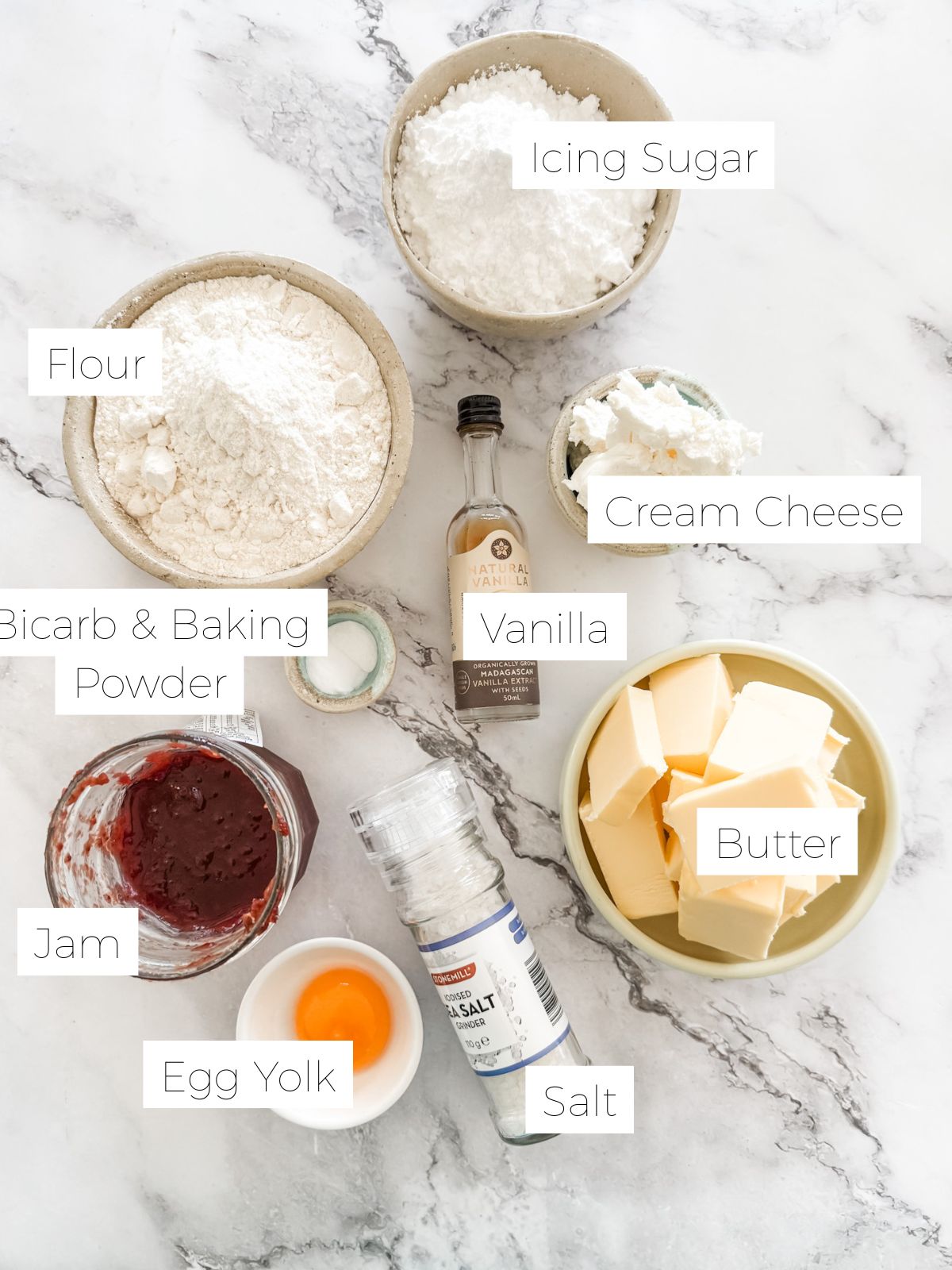 Ingredients in Strawberry Jam Drop Cookies - bowls of flour, icing sugar, butter, egg yolk, cream cheese & baking powder & vanilla bottle on white bench top. 