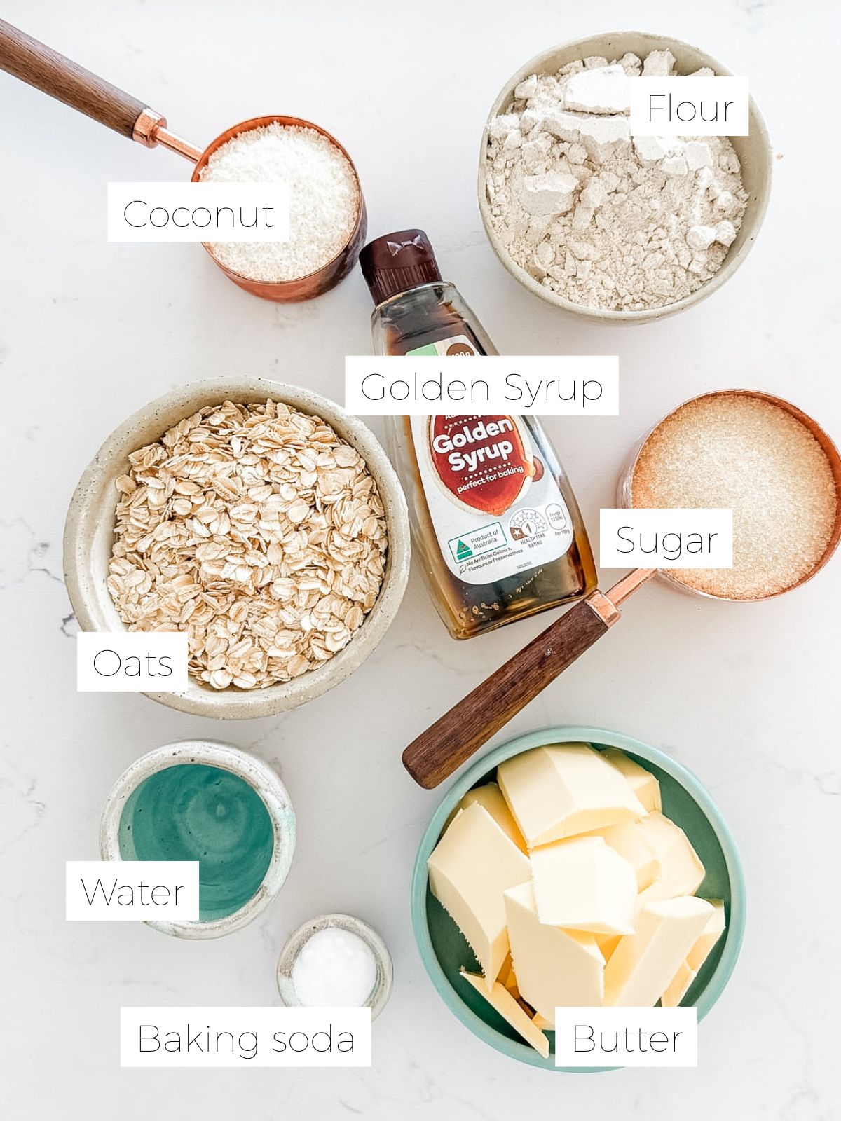 Ingredients in ANZAC biscuits - coconut, bowl of flour, bowl of oats, bowl of butter, measuring cup of sugar, bottle of golden syrup, small bowl of baking soda on white marble bench.