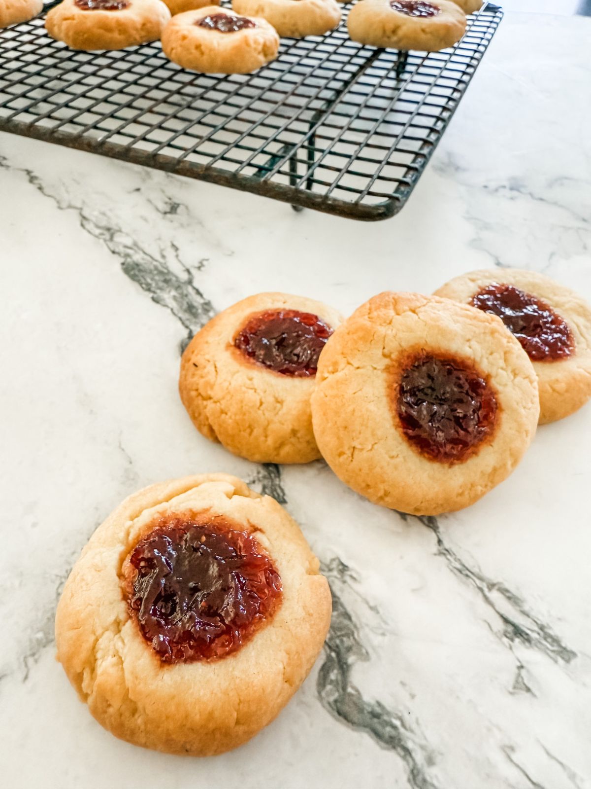 4 strawberry jam drop cookies on white marble bench with cooling rack in background. 