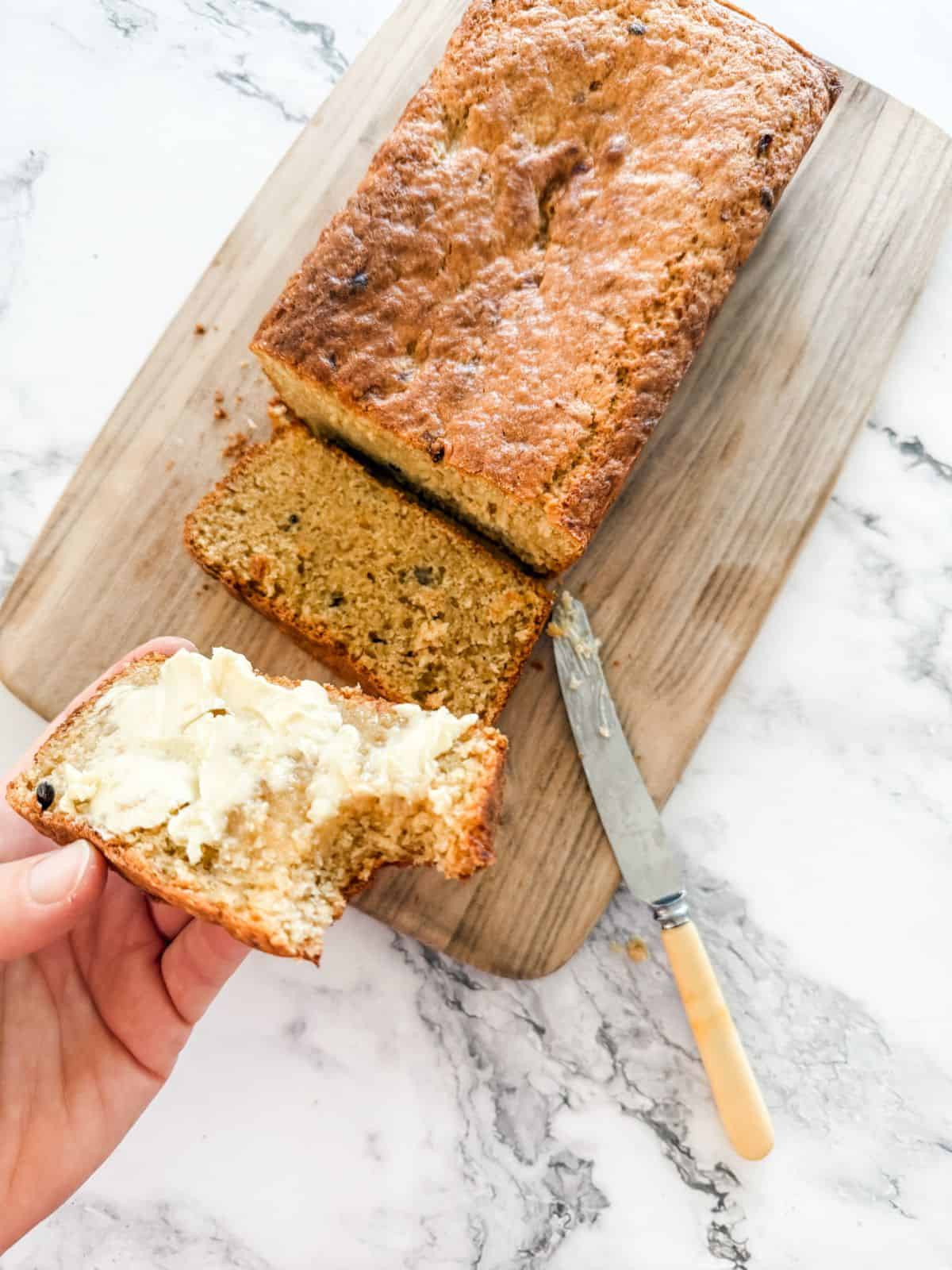 Slice of banana and passionfruit loaf spread with butter with a bite out held above full loaf. 