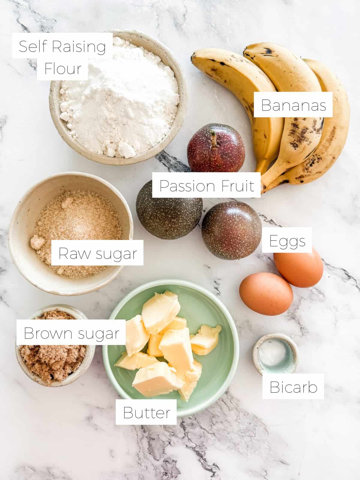 Ingredients in banana and passionfruit loaf - bowl of flour, sugar, butter, brown sugar - bananas, passionfruit & an egg on white bench top.