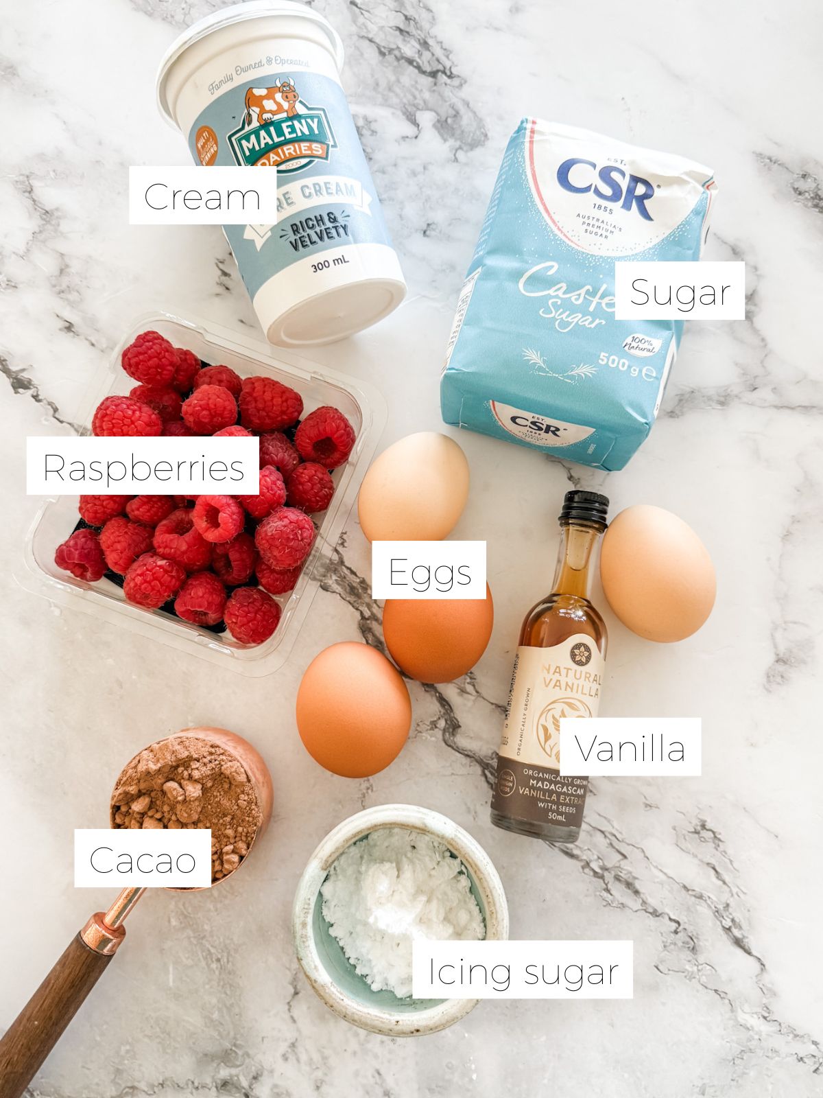 Tub of cream. Packet of sugar. Punnet of raspberries. 4 eggs. Bottle of vanilla. Measuring cup of cacao on white marble bench top to make chocolate and raspberry pavlova stack. 