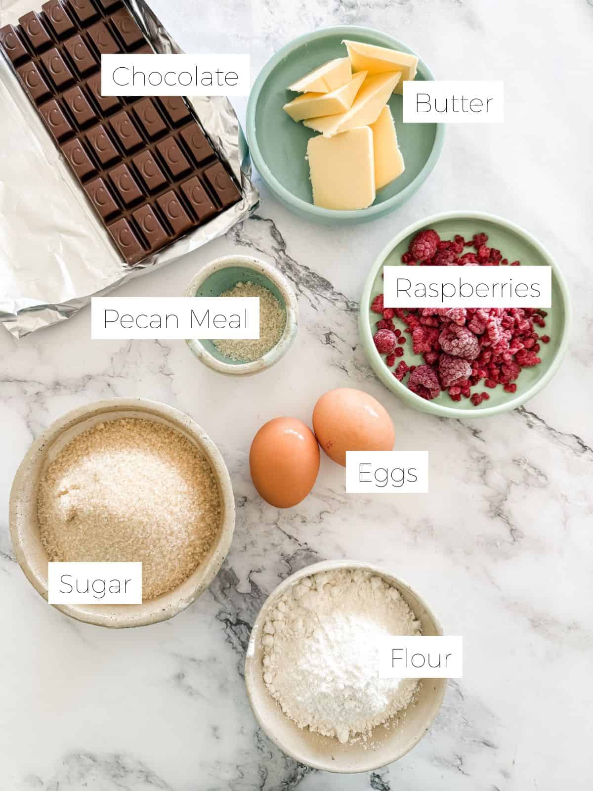 Ingredients in Raspberry and pecan brownies - block of chocolate, bowls of - butter, flour, sugar, raspberries & 2 eggs on white bench. 