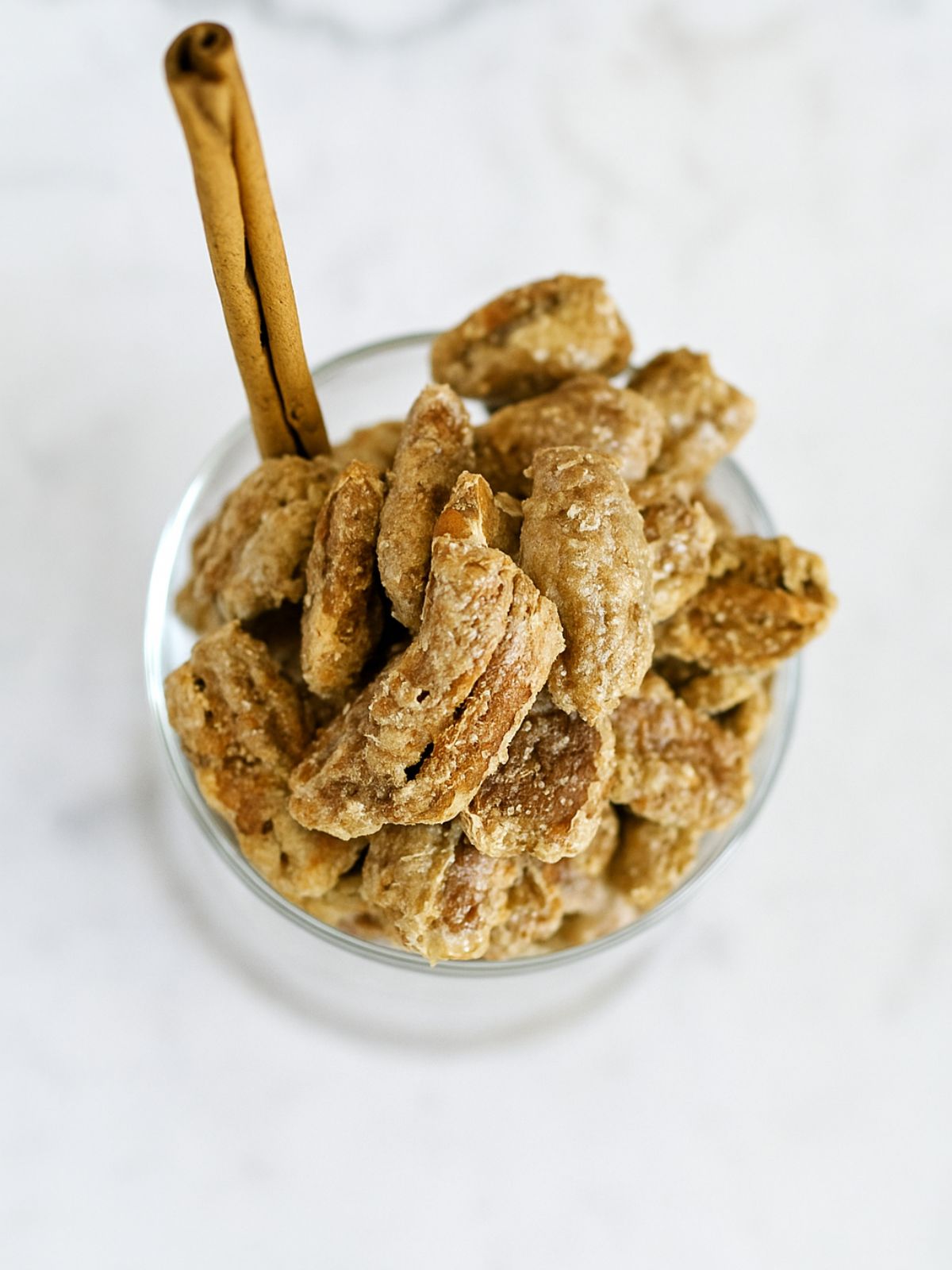 Sugared cinnamon pecans in a glass bowl with cinnamon quill on white marble bench top.