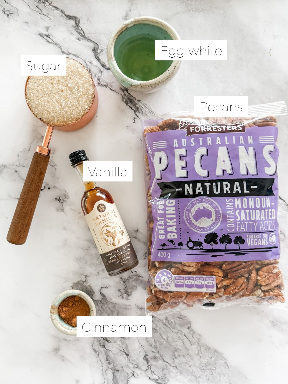 Bag of pecans, measuring cup of sugar, dish of cinnamon, bottle of vanilla and small bowl of egg white on white marble bench top.