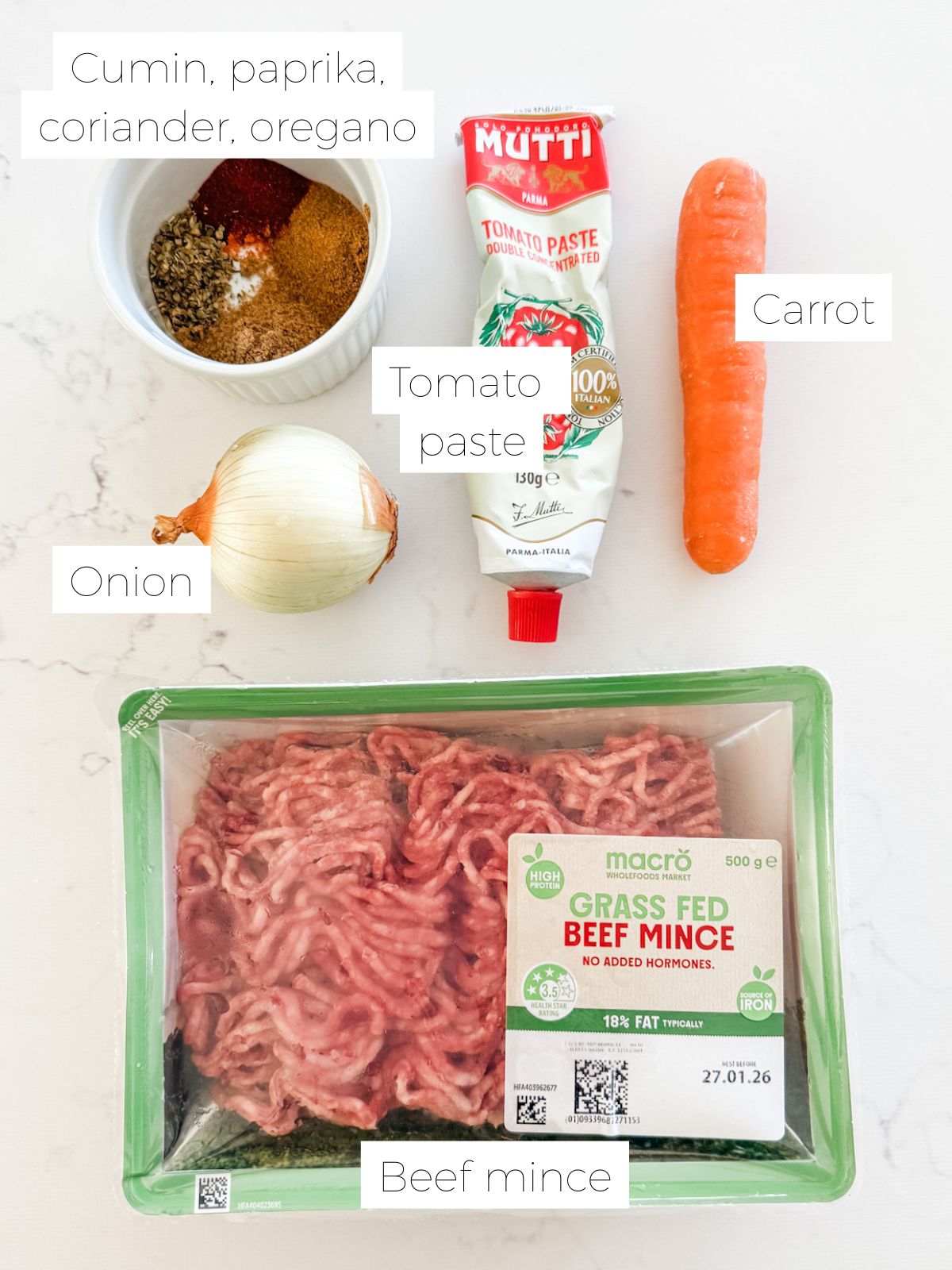 Packet of beef mince, Carrot. Tube of tomato paste. Onion & bowl of spices on a white marble bench. 