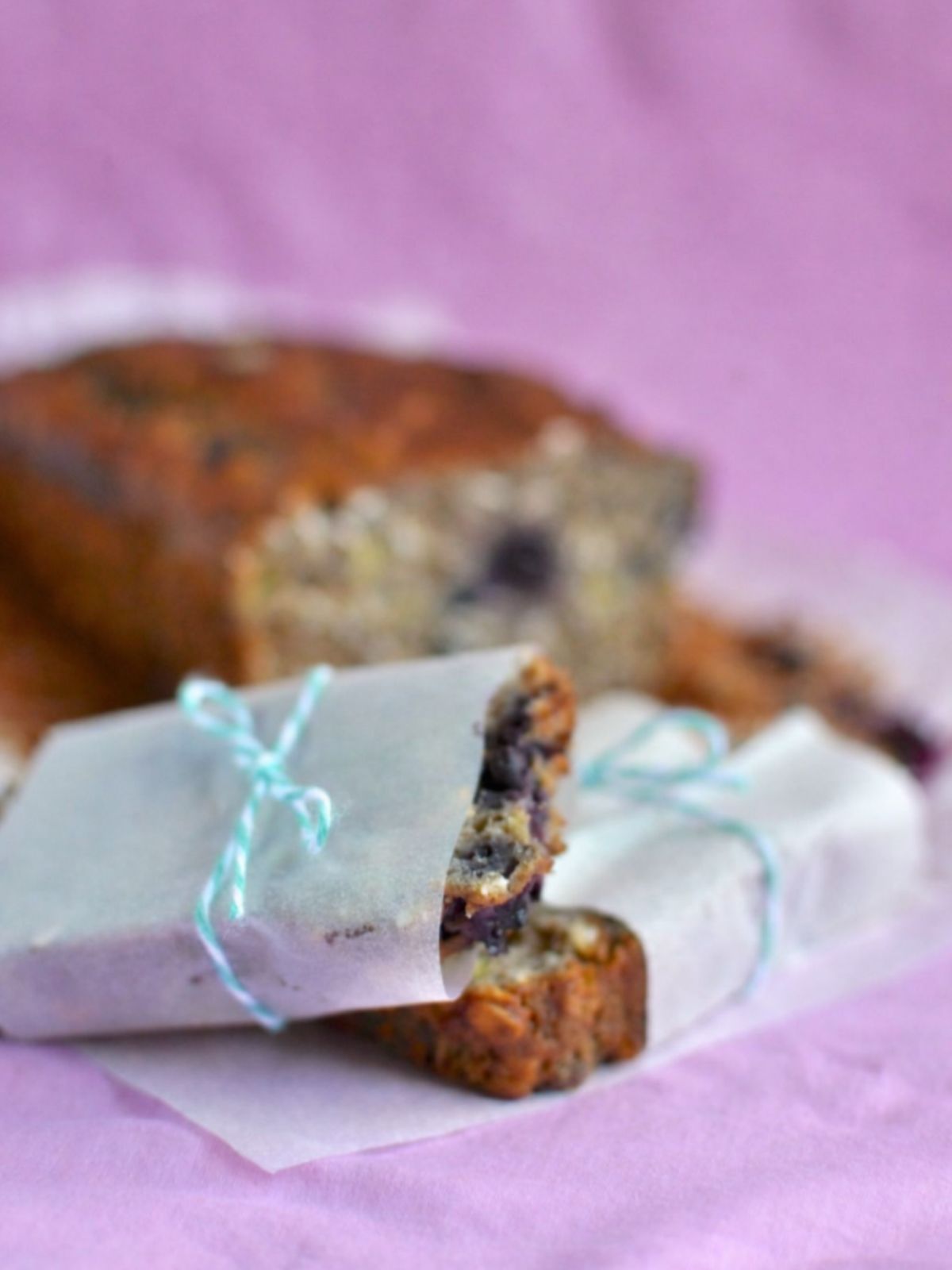 Loaf of banana bread in background with two slices wrapped in white parchment tied with string. 