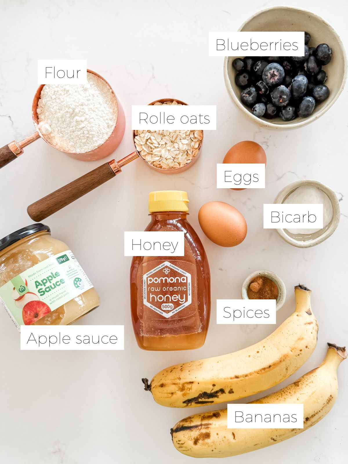 Honey, bananas, jar of apple sauce, bowl of blueberries, measuring cup of oats & flour and 2 eggs for making healthy banana bread with oats. 