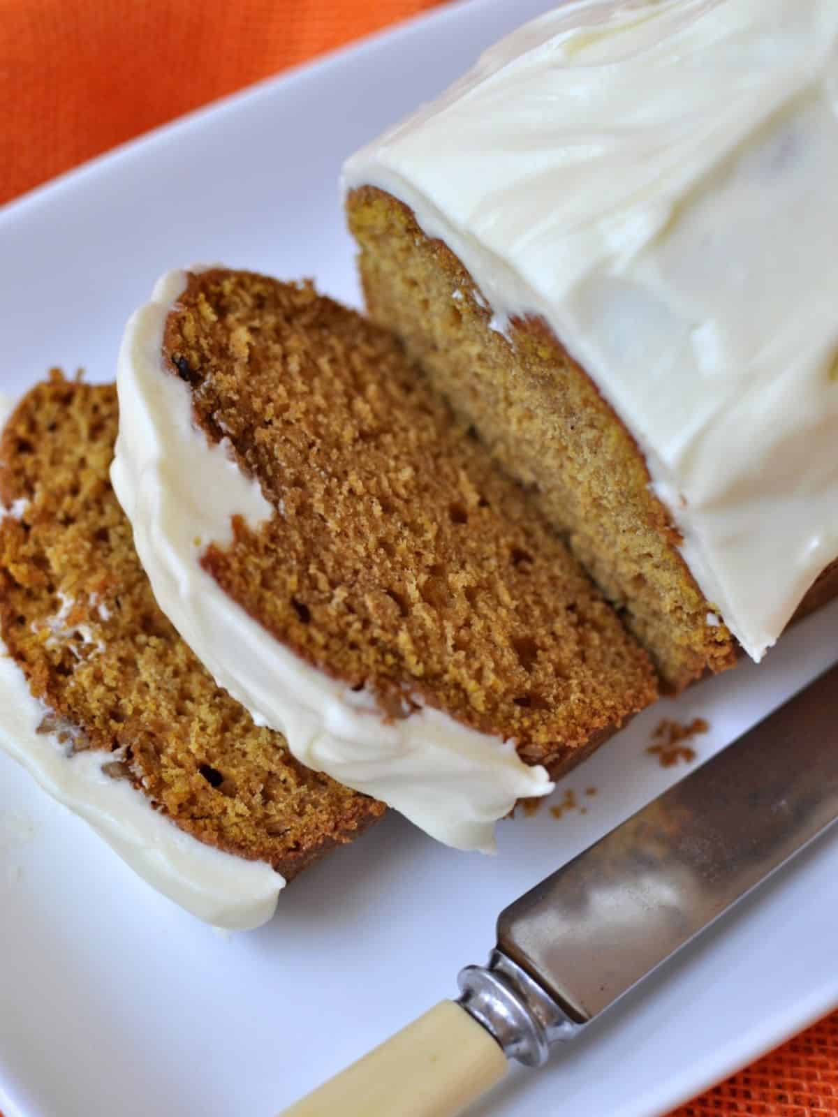 Pumpkin loaf cake with cream cheese icing with two slices cut and a bone butter knife on a white plate. 