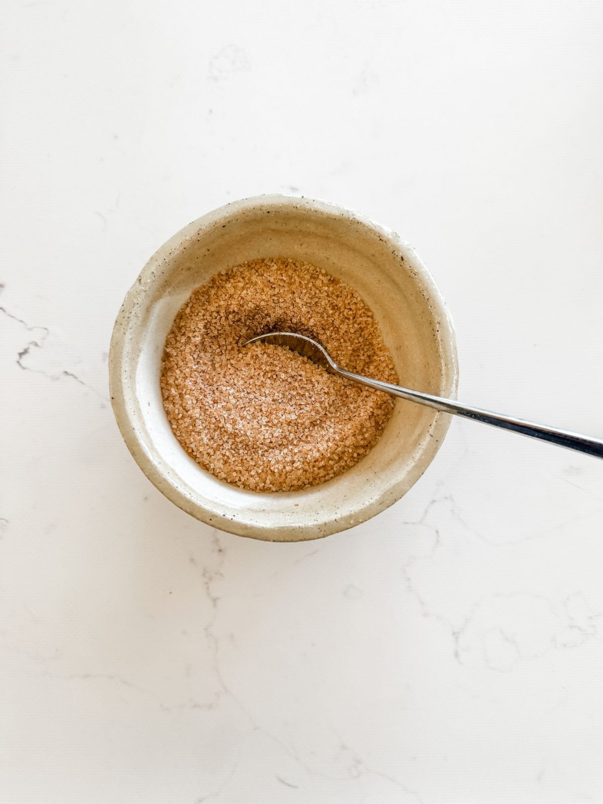 Cinnamon sugar in a beige bowl with a silver spoon on a white marble bench.