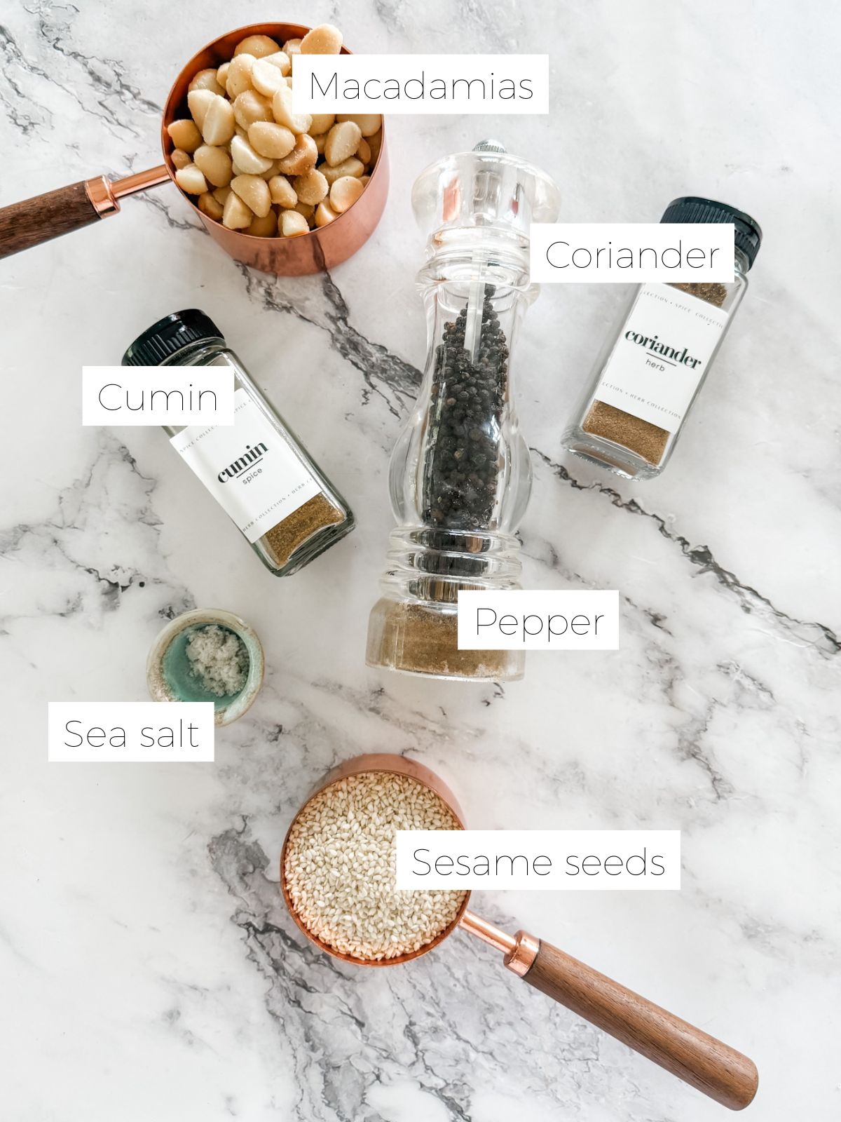 Measuring cups of macadamia nuts & sesame seeds, jar of coriander and cumin powder, pepper grinder and small bowl of sea salt for making macadamia dukkah on white marble bench top.