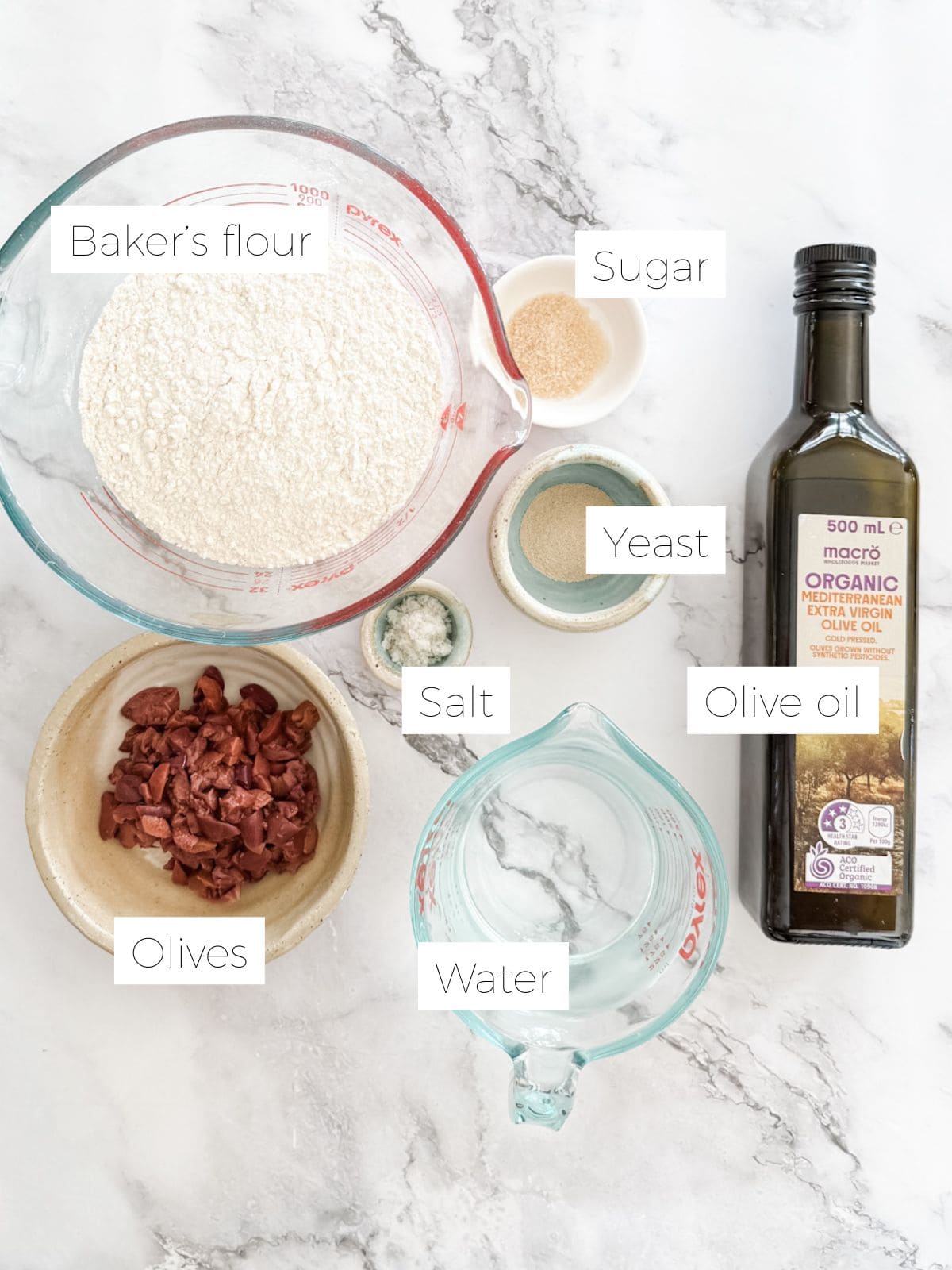 Ingredients laid out to make olive bread on a marble bench. Bread flour, yeast, salt, sugar, olive oil, water and black olives. 
