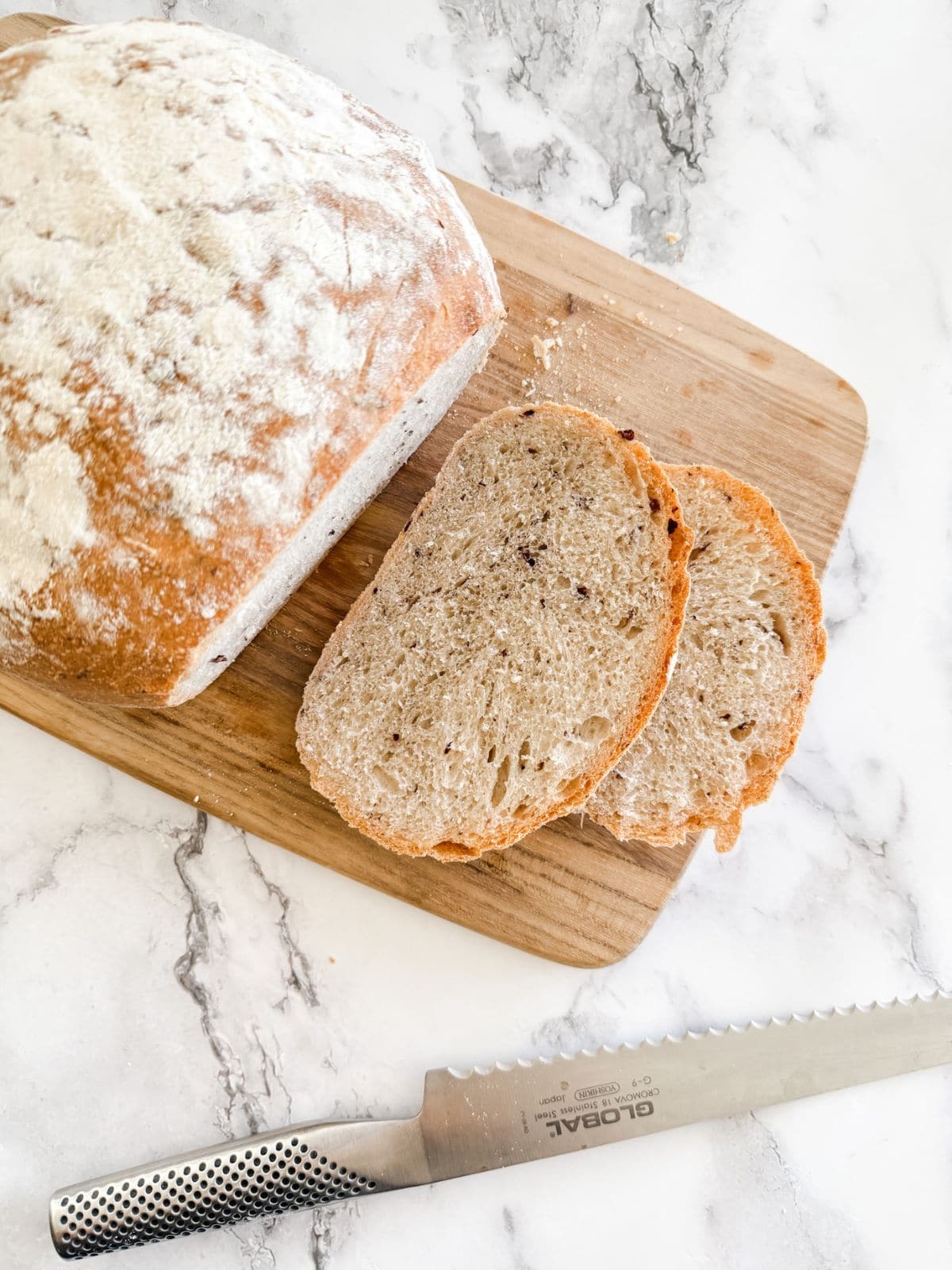 Olive bread with 2 slices out on a wooden chopping board with a silver bread knife on a bench top.