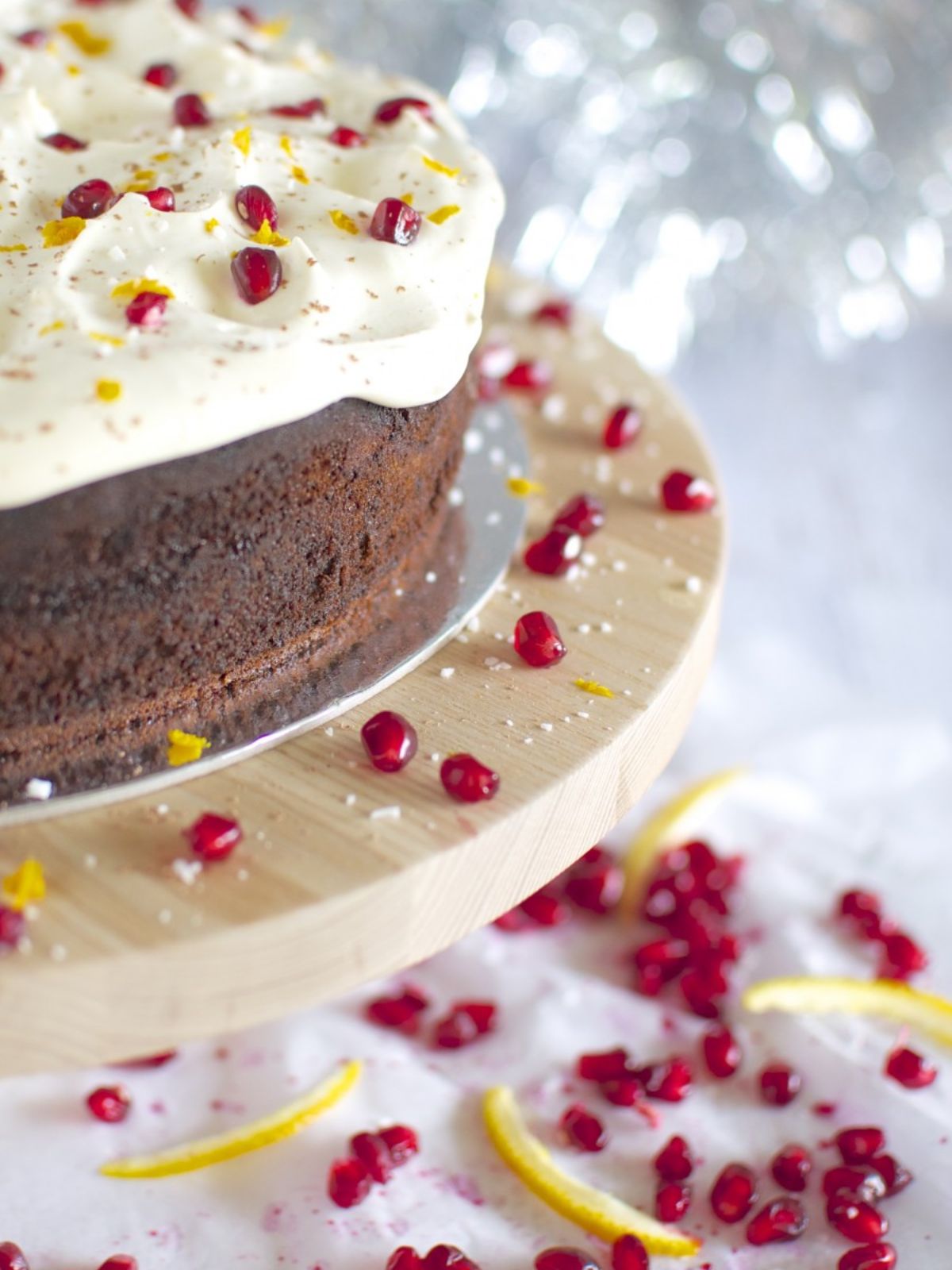 Chocolate cake on timber cake stand iced with white icing topped with pomegranate & orange zest.