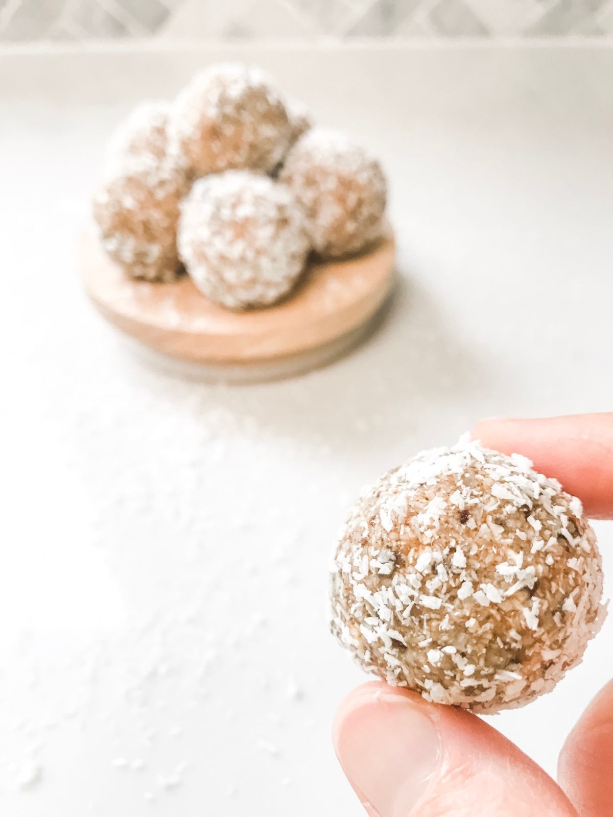 Banana bread bliss ball dusted with coconut held in two fingers on a white marble bench.