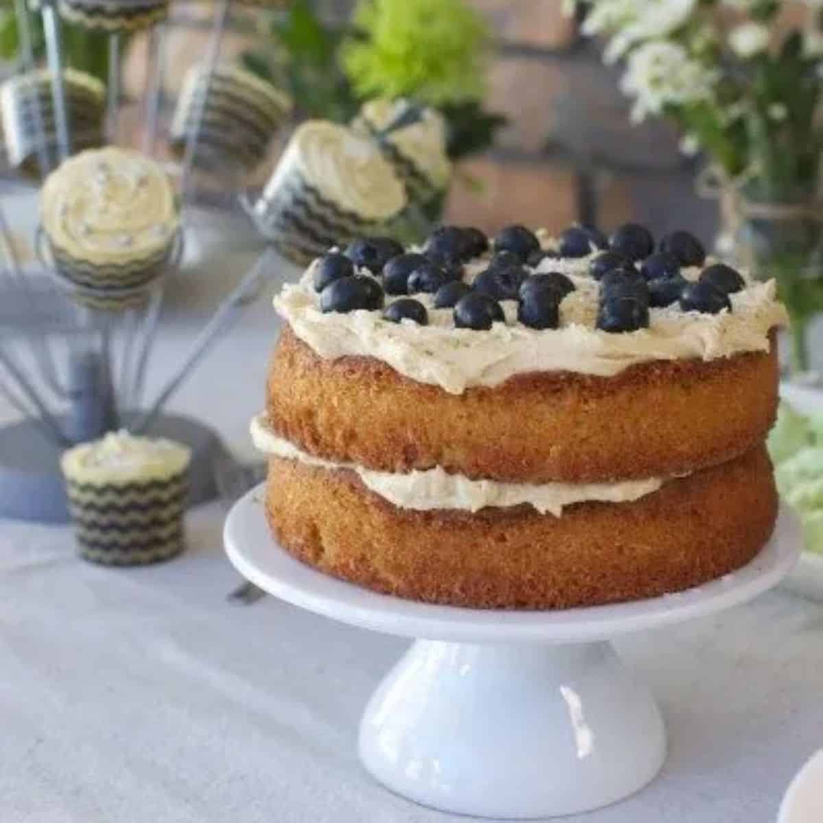 Gluten free lime and blueberry cake with coconut - double layer golden cake with white icing between layers and on top topped with fresh blueberries. Cake on a white cake stand.