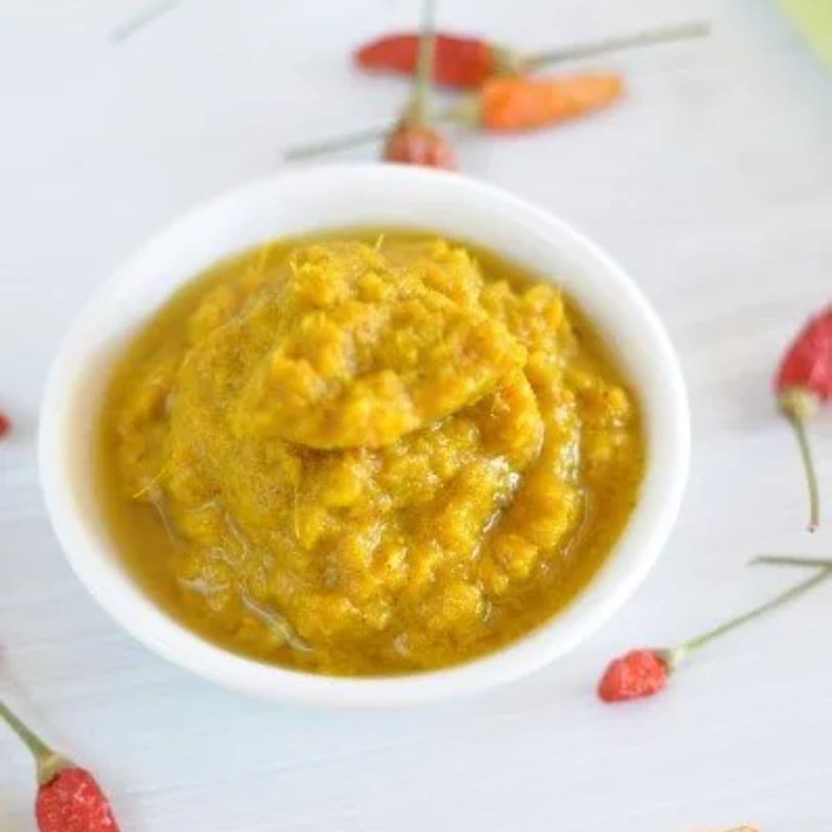 Thai yellow curry paste - blob of yellow curry paste in small white ramekin on white bench with fresh chilis around it.