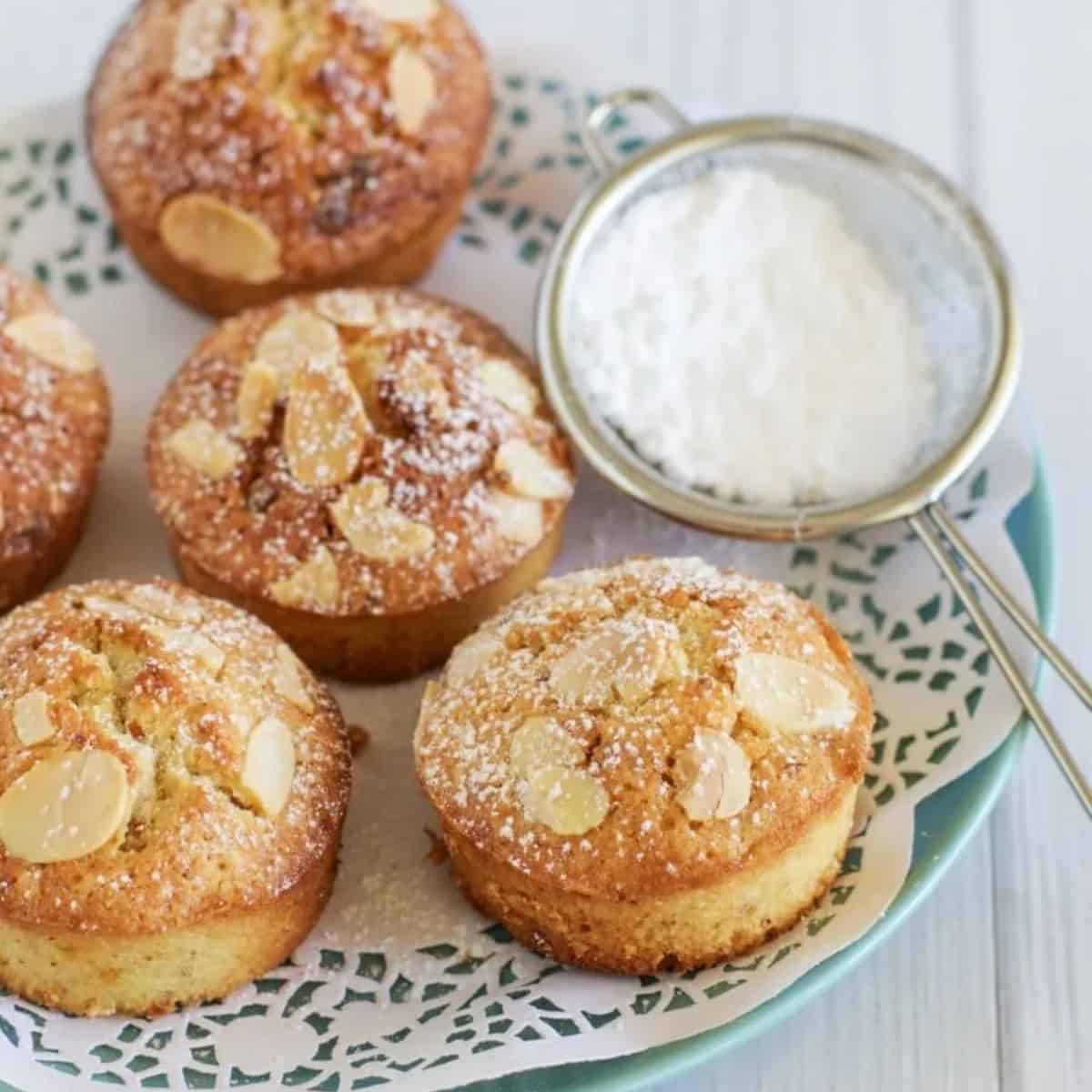 Passionfruit and white chocolate friands coated in flaked almonds. Arranged on a doilie on a plate with a sieve of icing sugar.