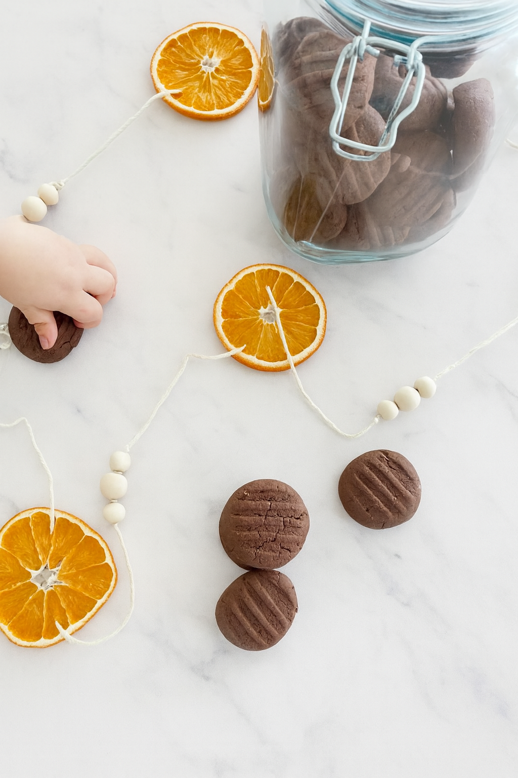 3 Chocolate Shortbread Cookies on white marble bench top with toddler hand and jar of cookies and dried orange garland. 