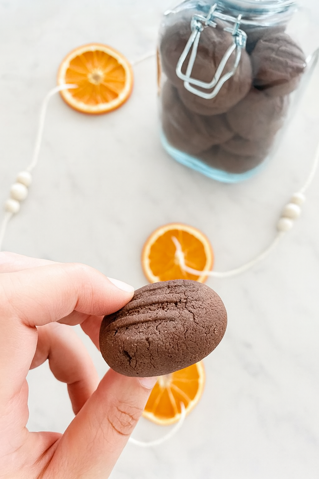 Hand holding Chocolate Shortbread Biscuit over white marble benchtop with dried oranges and jar of cookies on the bench. 