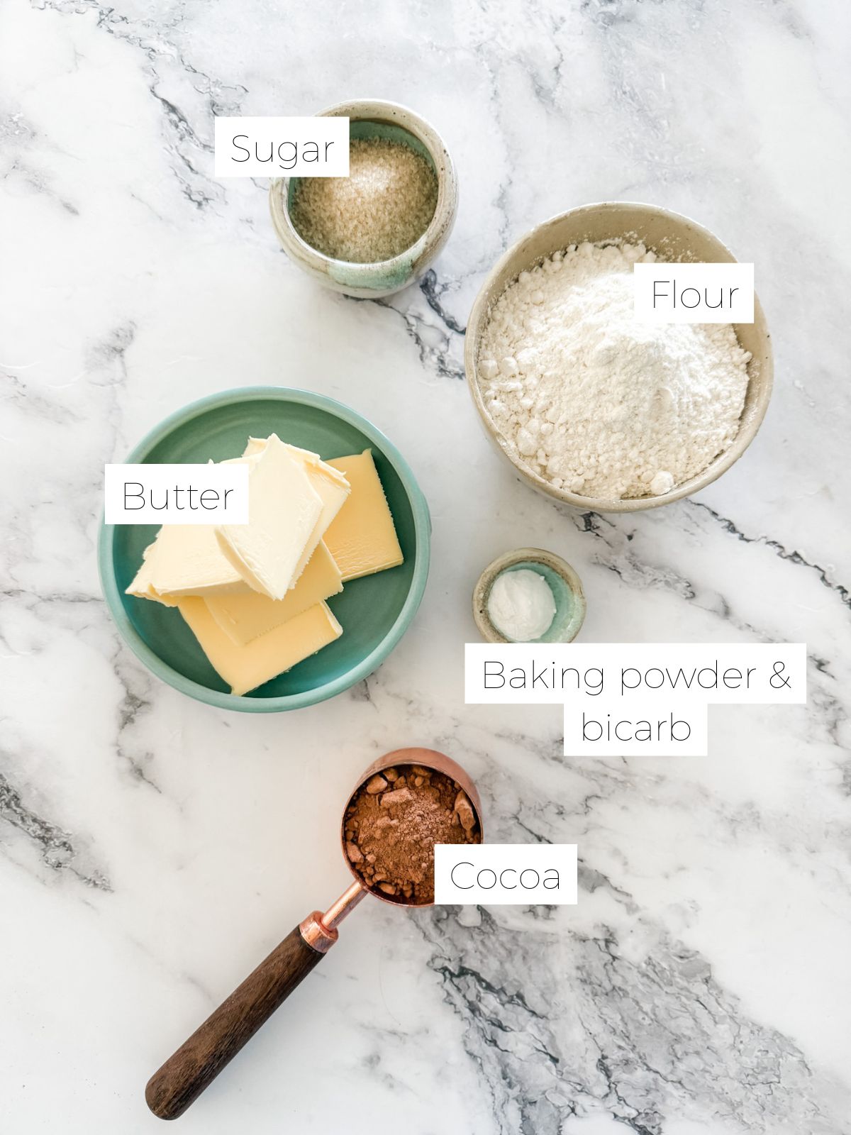 Ingredients in Chocolate Shortbread Cookies laid on white marble bench - in bowls - butter, flour, sugar, salt and measuring cup of cocoa. 