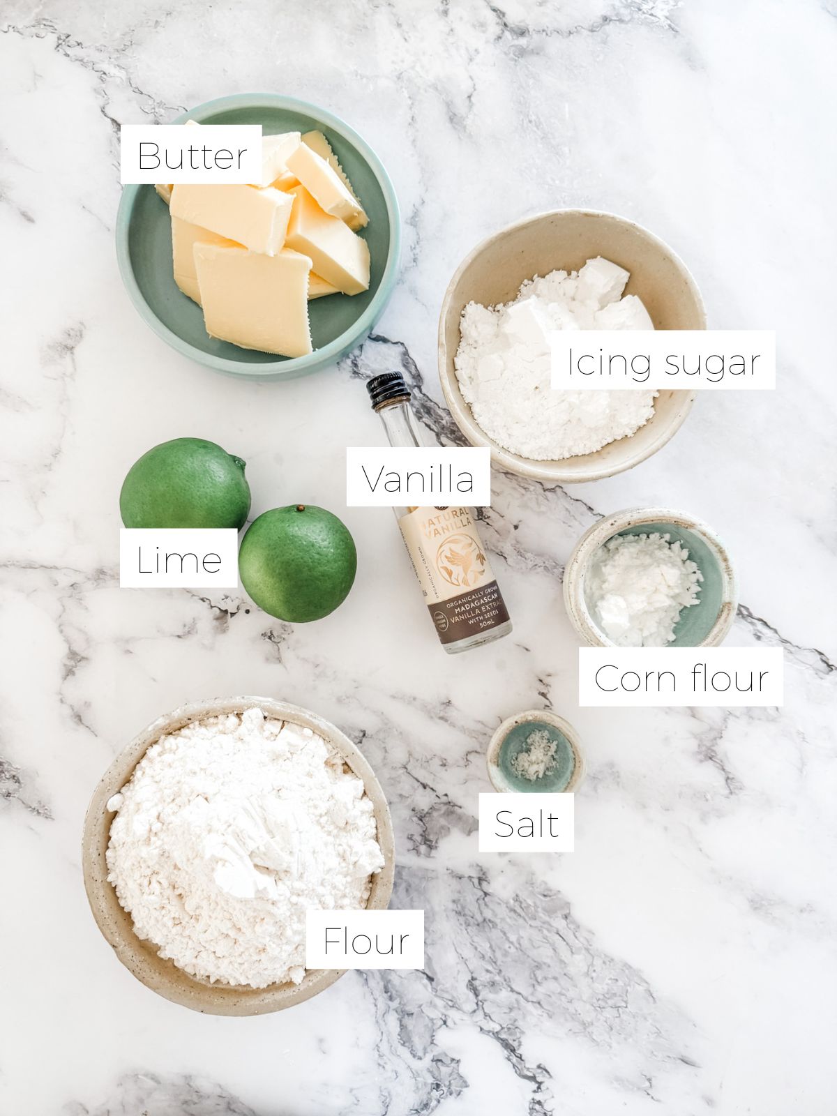 Bowl of butter, flour, icing sugar, corn flour, 2 limes, bottle of vanilla on white marble ready for malking lime meltaway shortbread biscuits. 