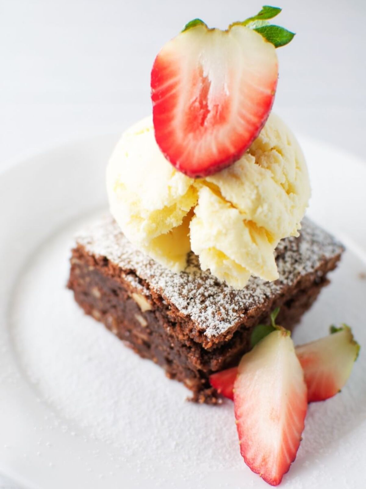 Square of chocolate brownie dusted with icing sugar topped with a scoop of ice-cream & strawberries on a white plate.