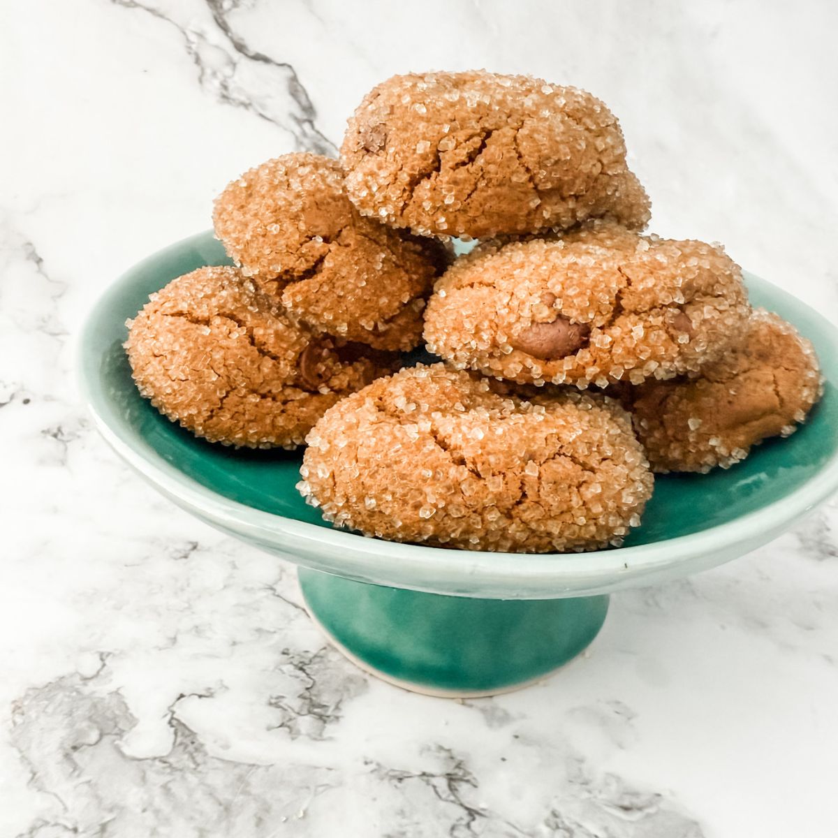 Pile of sugar crusted chocolate ginger cookies on a teal small cake stand on white marble bench top.