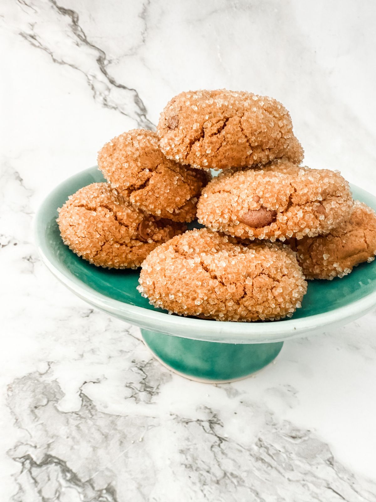 Pile of sugar crusted chocolate ginger cookies on a teal small cake stand on white marble bench top.