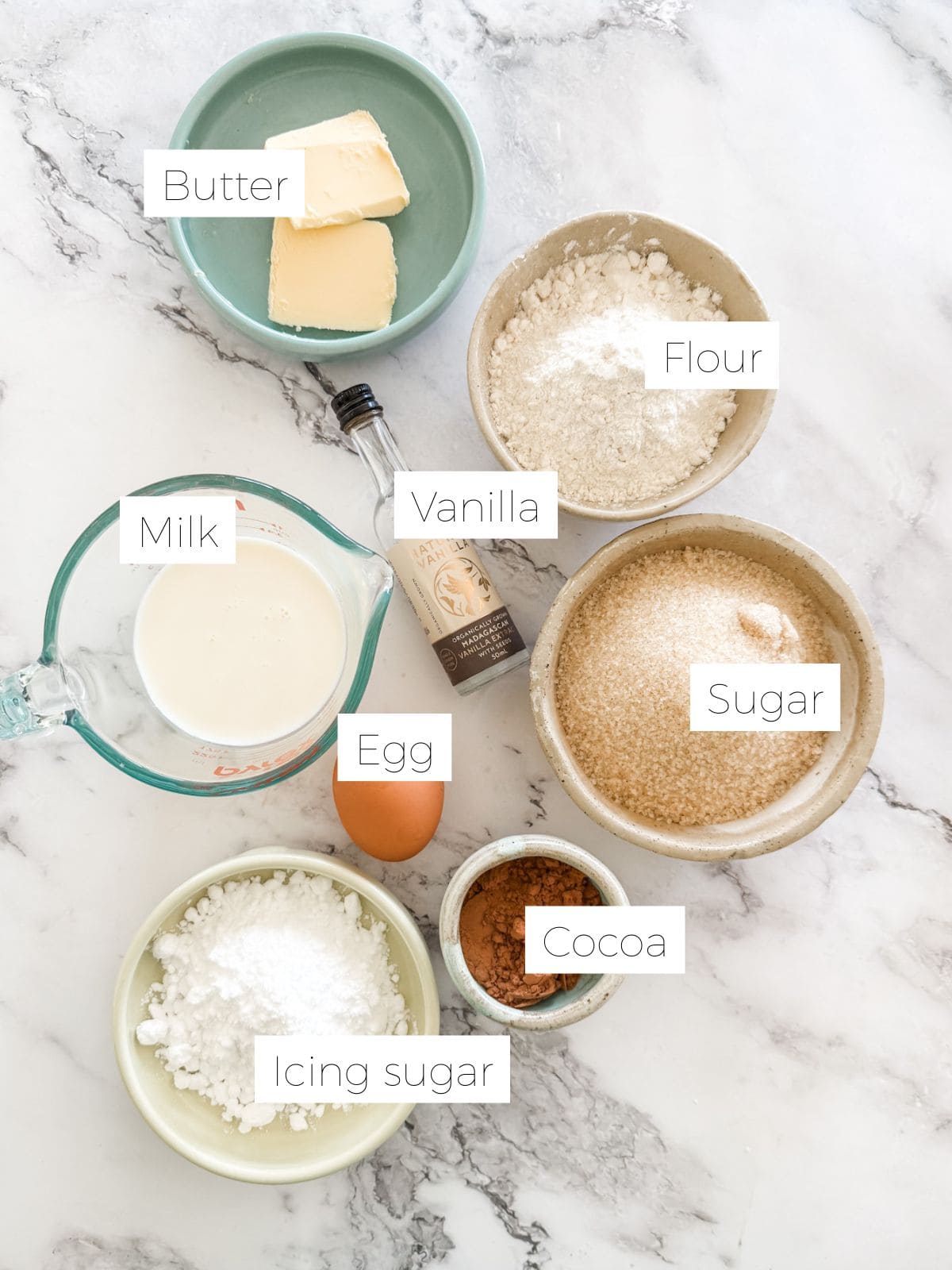 Bowls of butter, sugar, flour, icing sugar, cocoa. Jug of milk. Egg. Bottle of vanilla on white marble bench.