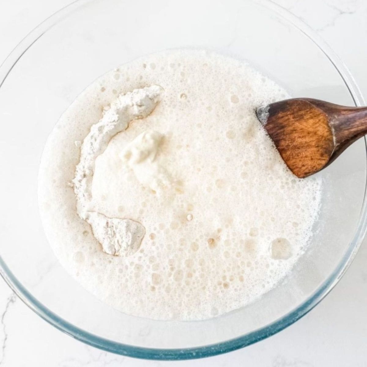Glass bowl with flour, lemonade & cream and a wooden spoon. On white marble bench.