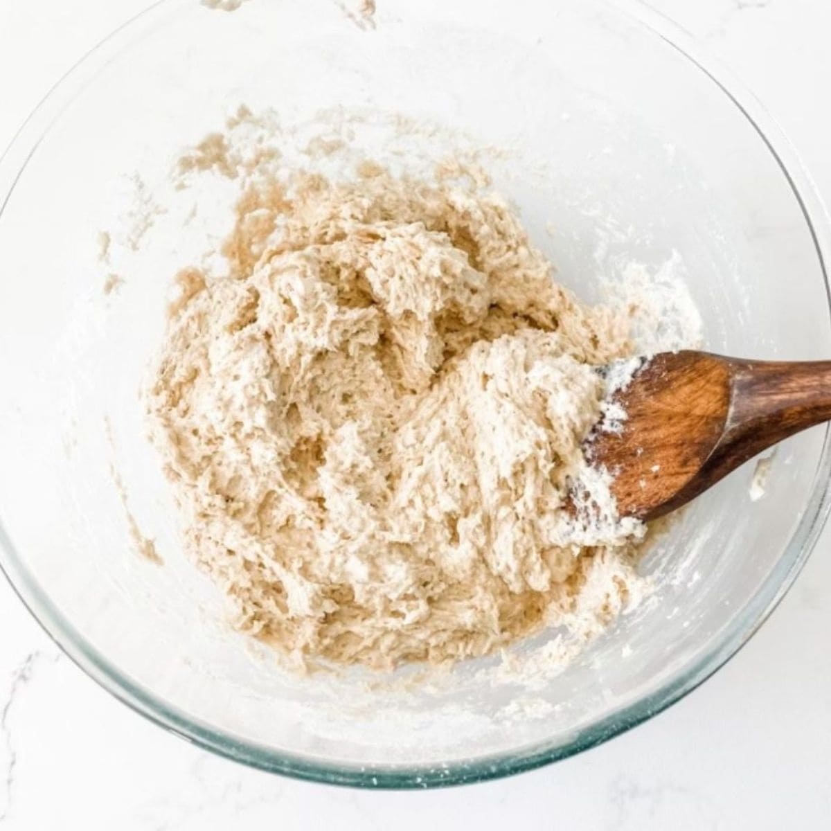 Scone dough in glass bowl with wooden spoon on white marble bench top.