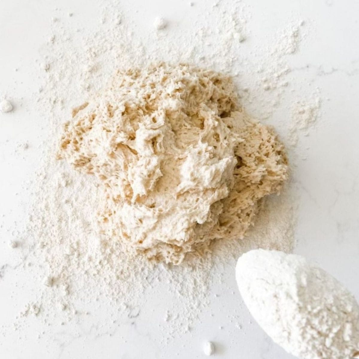 Scone dough in a heap on a white marble bench with spoon of flour.