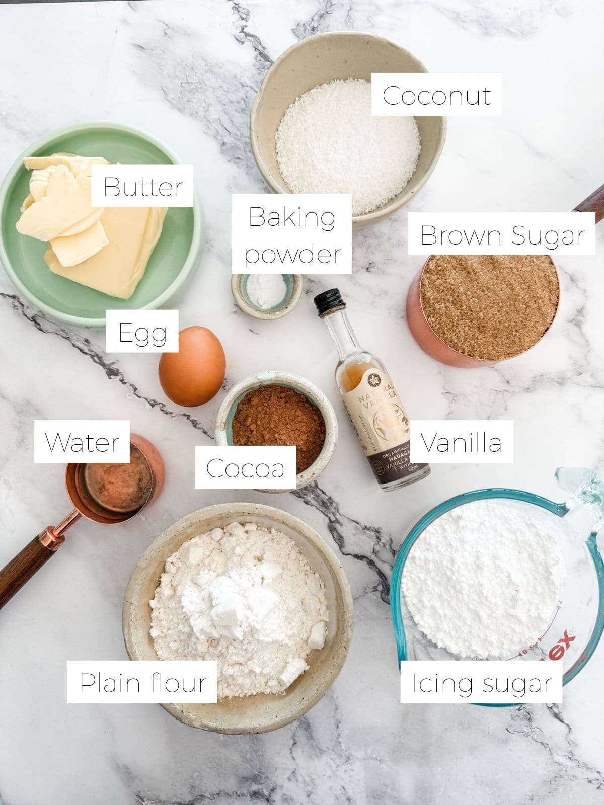 Ingredients in chewy chocolate slice. Individual bowls of butter, brown sugar, icing sugar, cocoa, coconut, flour, bottle of vanilla and an egg.