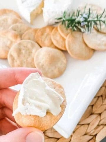 Homemade water cracker biscuit topped with white brie cheese in a white hand. In the background is a square white plate with more crackers and rosemary.