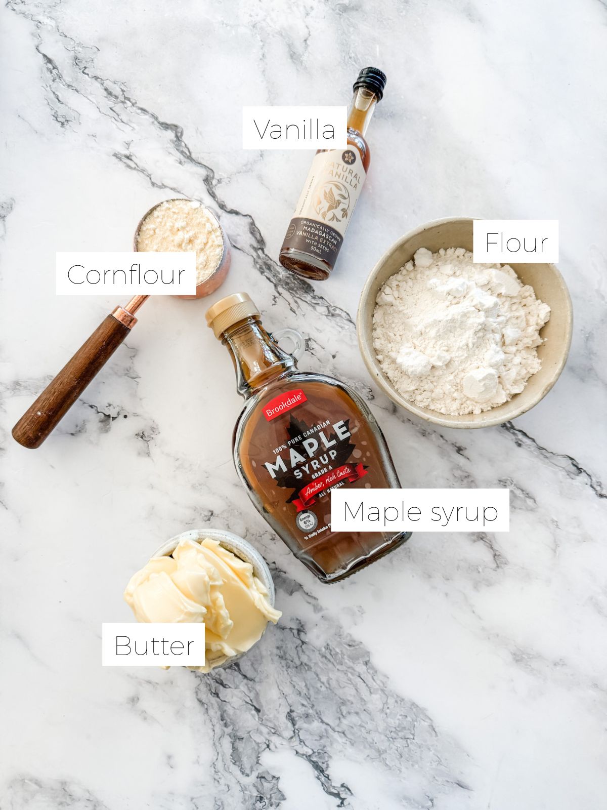 Ingredients in Maple Butter Biscuits - bowl of flour, copper measuring cup of cotn flour, bowl of butter, bottle of vanilla & maple syrup on white marble bench top. 