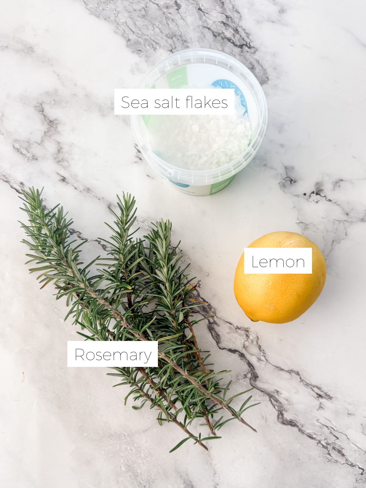 Sea salt flakes, fresh rosemary and a lemon on white marble benchtop ready to make lemon rosemary salt.