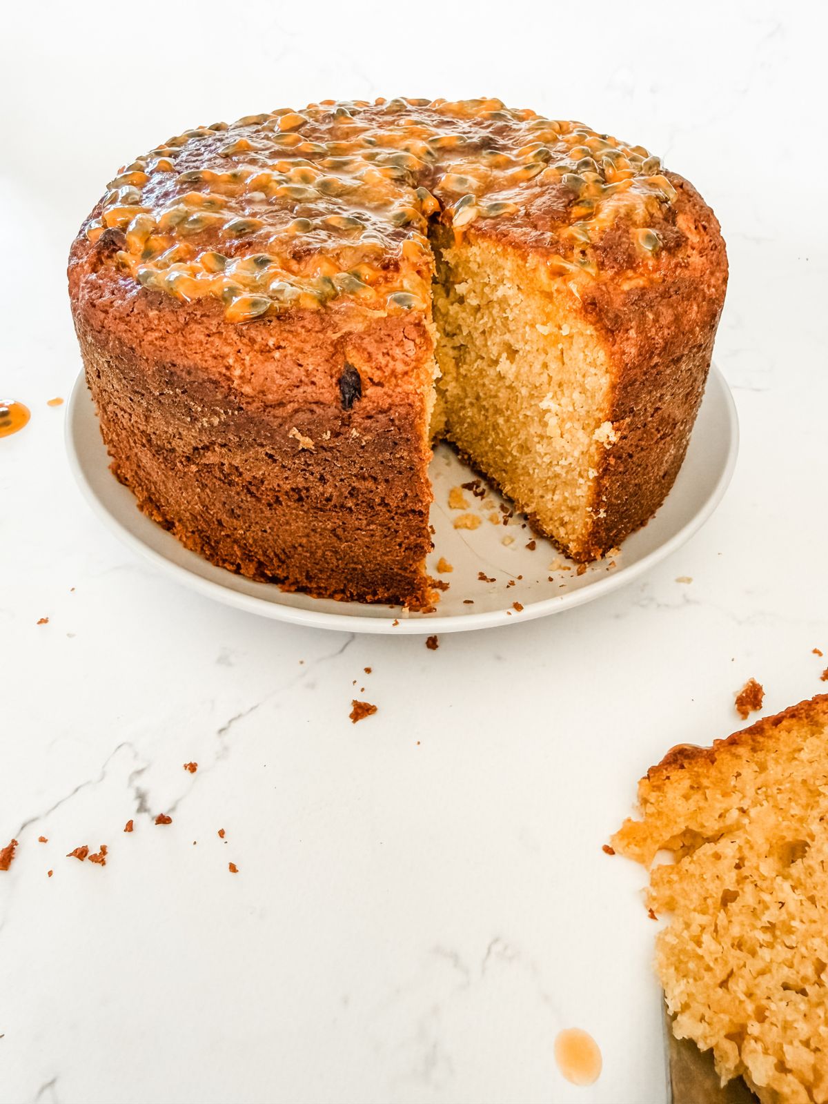 Easy Passionfruit Yoghurt Cake with slice cut out with a slice cut out of it on a white plate on white marble bench. 