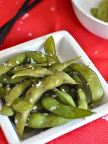 Japanese edamame in a white bowl sprinkled with sea salt on a red tablecloth.