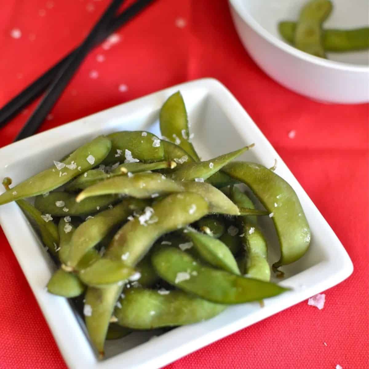Japanese edamame in a white bowl sprinkled with sea salt on a red tablecloth.