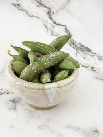 Small bowl of cooked edamame beans with salt on a white marble bench top.