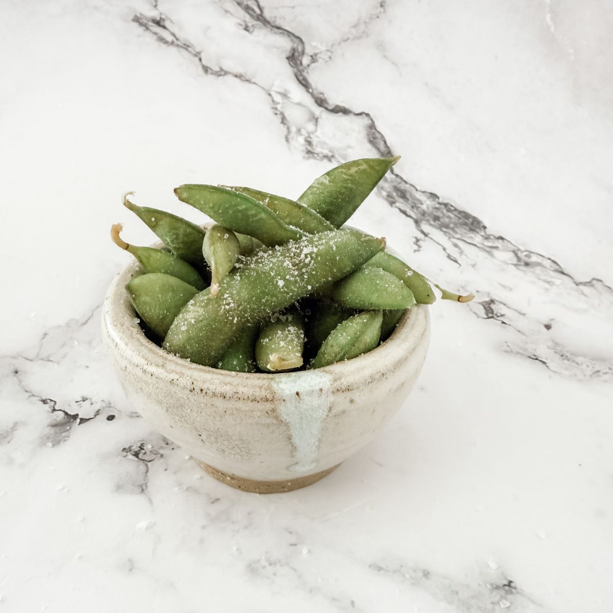 Small bowl of cooked edamame beans with salt on a white marble bench top.