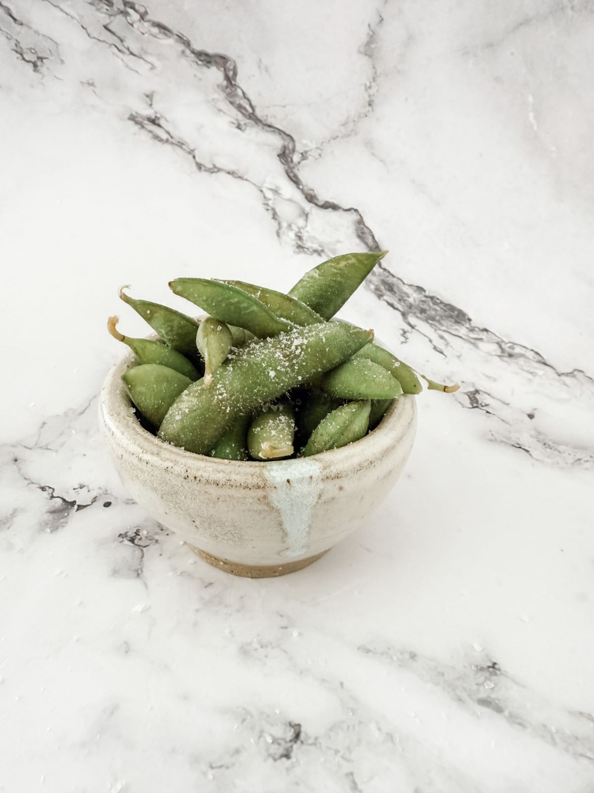 Cooked edamame beans in a small bowl with salt on a white marble bench top. 