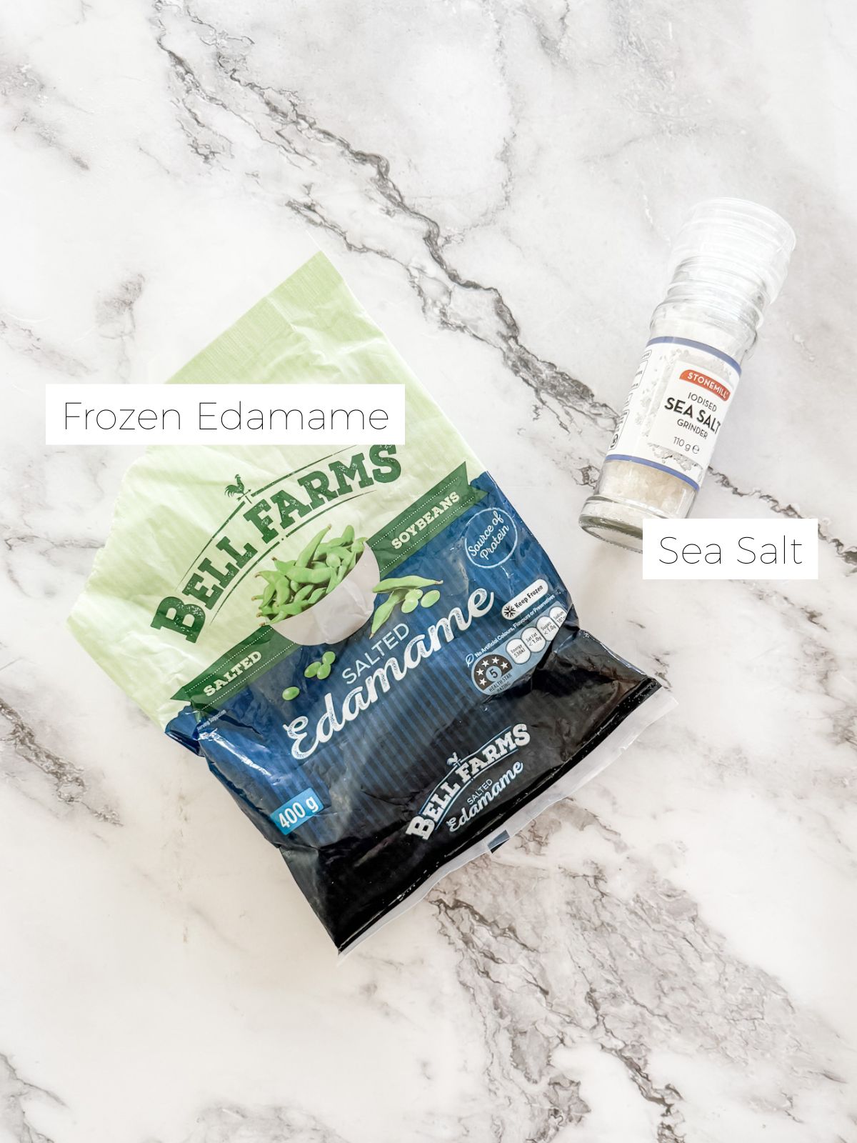 Packet of frozen edamame and salt shaker on white marble bench top. 
