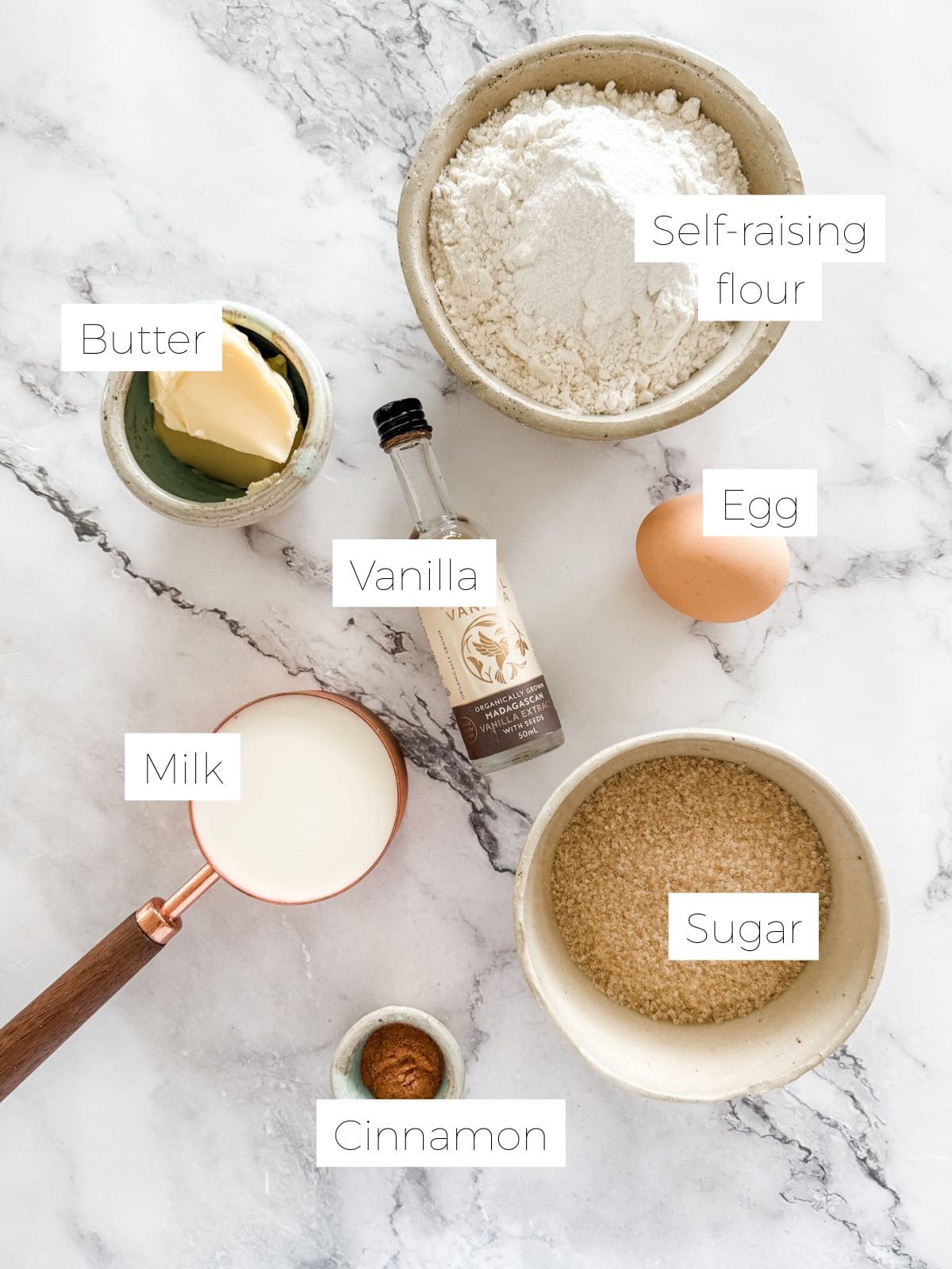 Bowl with flour, butter, sugar, cinnamon. Measuring cup with milk. Small bottle of vanilla. An egg. All on white marble benchtop.