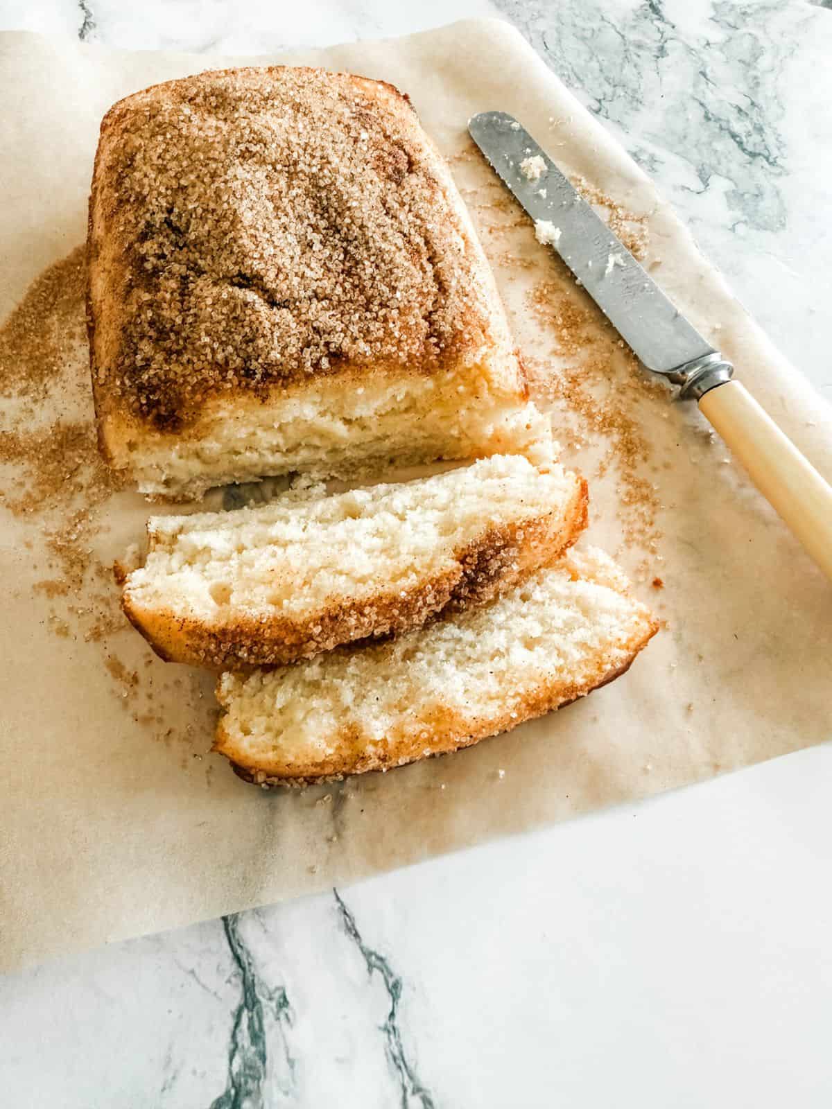Cinnamon tea cake with two slices out on a sheet of baking paper on a white bench with a bone butter knife.