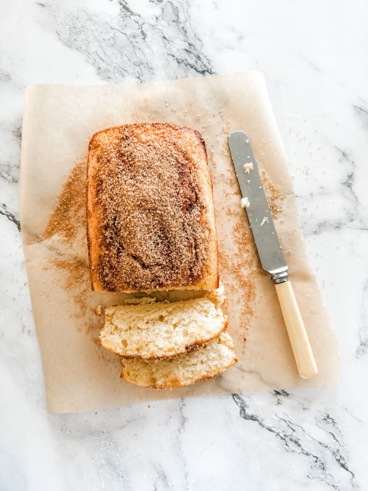 Cinnamon tea cake with two slices out on a sheet of baking paper on a white bench with a bone butter knife.